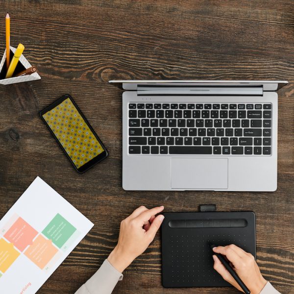 Overview of hands of contemporary creative designer sitting by wooden table in front of laptop and using stylus and graphic tablet